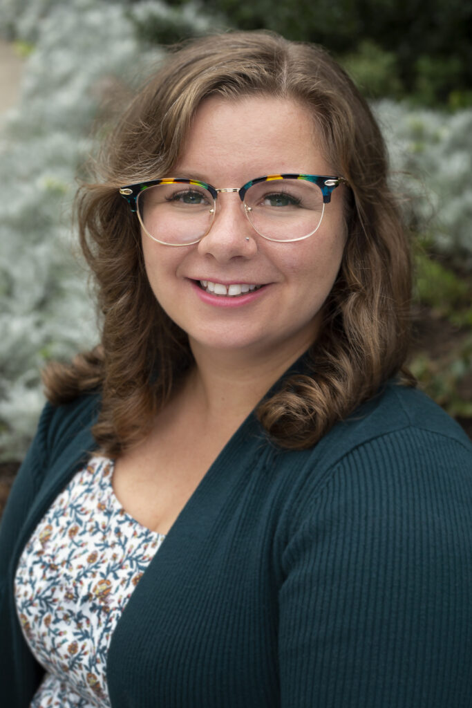 Image of Hayley Stefan. She is a smiling white woman with shoulder-length light brown curly hair, wearing a dark green blazer over a white dress with dark green vines and orange flowers and gold-rimmed glasses with a green, blue, and yellow border. She is seated and looks at the camera diagonally across her shoulder in front of a blurry background of plants.